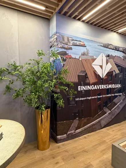 Grænar greinar í háum gylltum vasa við vegg með mynd og merki Einingaverksmiðjunnar
