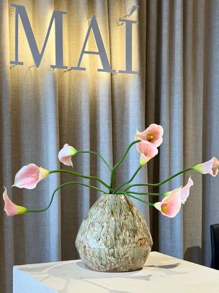 Upplýst MAÍ-merki á vegg og bleikar kallaliljur í rifflaðum vasa fyrir framan gardínur