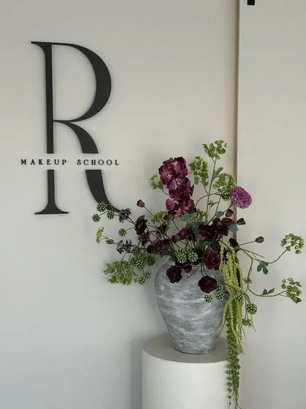 R Makeup School-merki á vegg og dökkrauð skreyting í gráum steinvasa á hvítum stalli